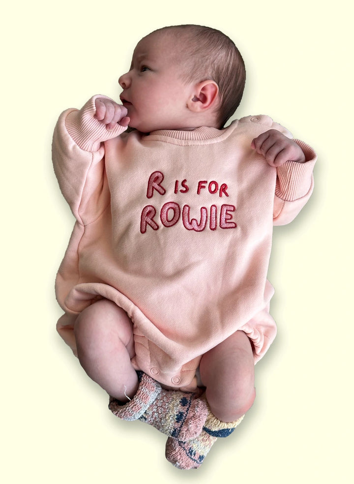 Baby wearing a pink onesie with 'R is for Rowie' text on a light background